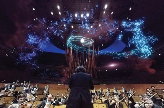 Image of a male conductor standing in front of an orchestra on a raised platform with a blue colored light display shimmering in the air above their heads.