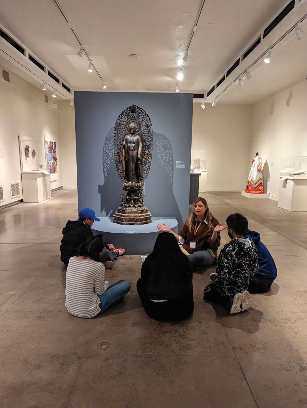 A gallery guide sits on the floor in an exhibition with a group of children scattered around listening.