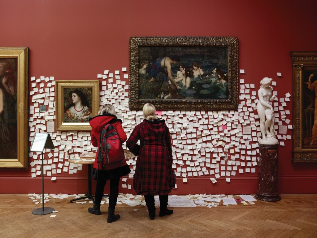 Two women stand in front of two ornately framed pieces of art, surrounded by white Post-It notes.