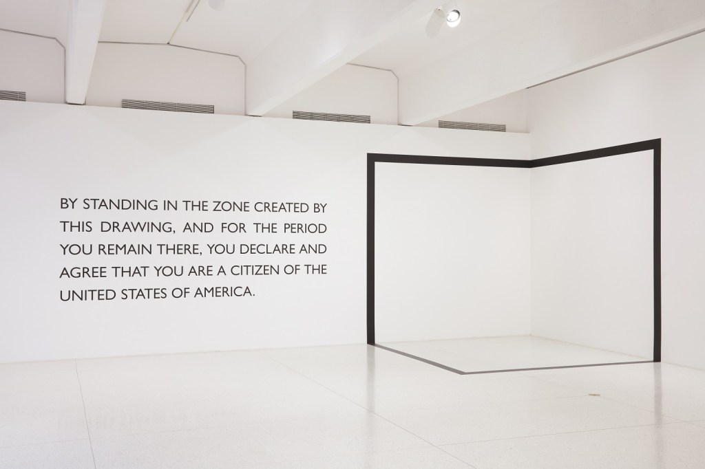 A white wall with a black box sectioned out has the following saying on it, "By standing in the zone created by this drawing, and for the period you ramain there, you declare and agree that you are a citizen of the United States of America." 