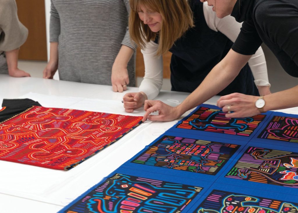 Several people lean over a table investigating two pieces of textile. 